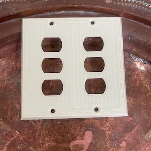 May include: Off-white, double-gang electrical outlet cover with six openings. The cover has a textured, vertical-line pattern and a decorative border. The cover is made of a solid, opaque material.