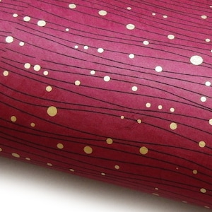 Op de afbeelding: Close-up van een rol bordeauxrood papier met een patroon van zwarte golvende lijnen en goudkleurige stippen. Het papier heeft een textuur en is gedeeltelijk opgerold, waardoor een witte rand zichtbaar is.