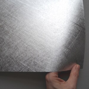 May include: A sheet of silver-colored metal with a textured, brushed appearance. The sheet is being held by a hand, revealing its curved edge. The metal has a reflective quality, catching the light and displaying a subtle sheen.