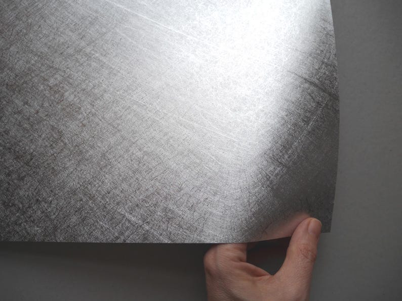 May include: A sheet of silver-colored metal with a textured, brushed appearance. The sheet is being held by a hand, revealing its curved edge. The metal has a reflective quality, catching the light and displaying a subtle sheen.