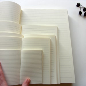 Puede incluir: Varios cuadernos rayados de diferentes tamaños, con páginas de color crema. Los cuadernos están abiertos, mostrando las páginas interiores con líneas horizontales. Una mano sostiene uno de los cuadernos más pequeños. Una ramita de bayas negras en la esquina superior derecha.