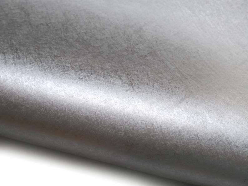 May include: Close-up of a roll of silver-colored fabric. The material has a textured surface with subtle variations in tone, creating a metallic sheen. The fabric is slightly curved, revealing its flexible nature.