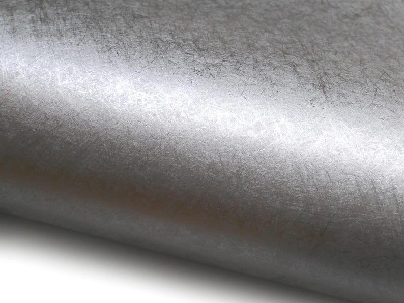 May include: Close-up of a textured, metallic silver surface. The material has a brushed appearance with subtle variations in tone and visible fine lines, creating a sense of depth and a tactile quality. The surface is slightly curved.