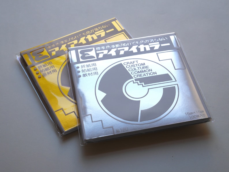 May include: Two packs of origami paper, one silver and one yellow. The silver pack has the text "CRAFT CUSTOM CULTURE COMMON CREATION" and "15cm x 15cm 100枚入り". The yellow pack has the text "アイアイカラー" and "折紙用 貼絵用 教材用".