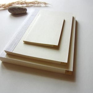 Puede incluir: Tres cuadernos de color crema apilados uno encima del otro. El cuaderno de arriba tiene una encuadernación blanca con un patrón texturizado. El cuaderno de abajo tiene una encuadernación de color crema con el número "2" impreso en él.