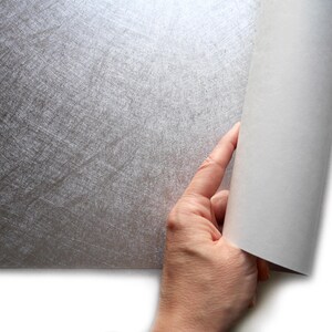 May include: A hand is shown rolling back a sheet of silver-colored paper, revealing a white interior. The silver paper has a textured, scratched appearance. The paper is likely a decorative or craft material.