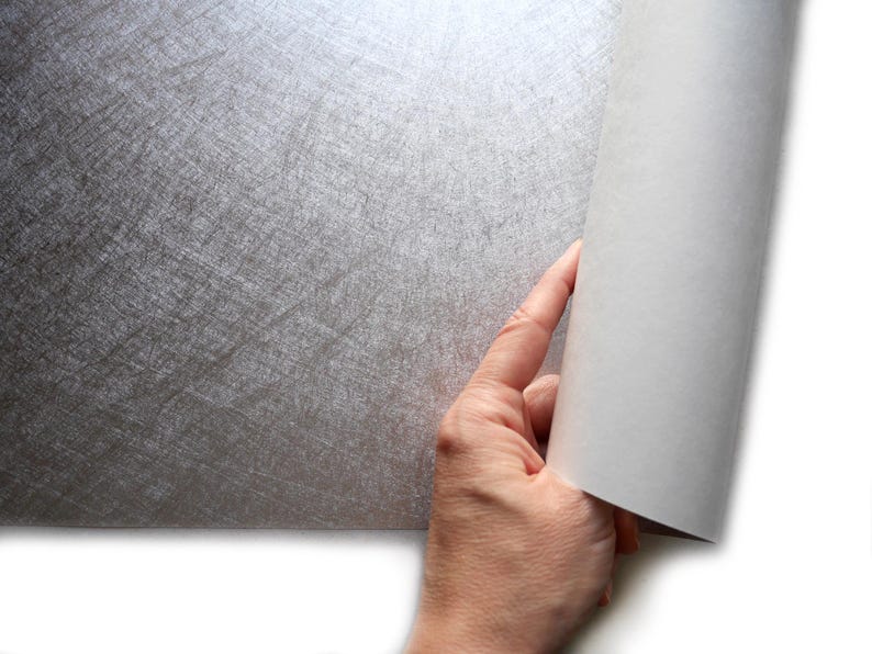 May include: A hand is shown rolling back a sheet of silver-colored paper, revealing a white interior. The silver paper has a textured, scratched appearance. The paper is likely a decorative or craft material.