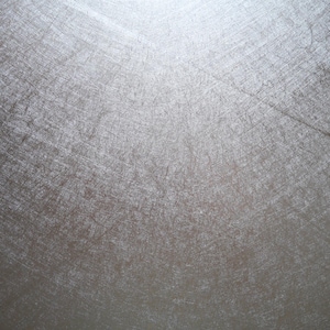 May include: A close-up shot of a textured, metallic surface. The surface has a silver-gray color with visible scratches and imperfections, creating a weathered appearance. The light source is at the top, casting shadows and highlighting the texture.