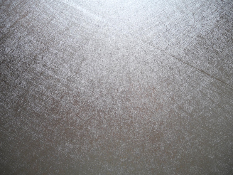 May include: A close-up shot of a textured, metallic surface. The surface has a silver-gray color with visible scratches and imperfections, creating a weathered appearance. The light source is at the top, casting shadows and highlighting the texture.