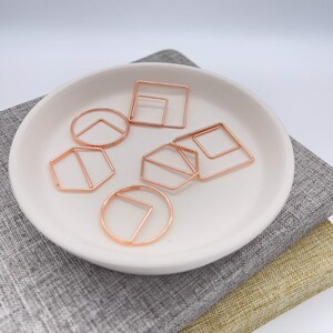 May include: A collection of rose gold geometric paper clips in various shapes, including squares, circles, and pentagons, arranged on a white ceramic dish. The dish rests on a gray and gold textured fabric.