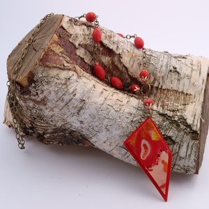 Peut inclure: Un collier avec un pendentif géométrique rouge et orange, une chaîne et des perles rouges. Le collier est présenté sur un morceau de bois de bouleau.