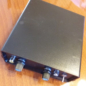 Könnte beinhalten: Schwarzes elektronisches Gerät mit zwei Knöpfen und einem Netzschalter. Das Gerät hat ein schwarzes Kunststoffgehäuse und einen einzigen RCA-Stecker.