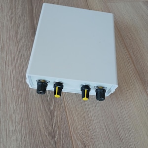 May include: A white rectangular electronic device with four black and yellow knobs. The device is on a wooden surface. The knobs are evenly spaced along the bottom edge of the device.
