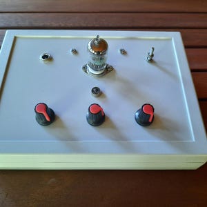 Può includere: Una scatola bianca con un tubo a vuoto argentato, tre manopole nere con cime rosse e un jack argentato sul davanti.