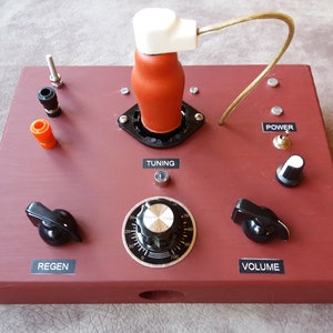 May include: A red wooden box with a vintage radio receiver. The receiver has a tuning knob, a volume knob, a regen knob, and a power switch. The receiver also has a red and white vacuum tube with a white ceramic insulator.