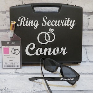 May include: Black ring security case with the text "Ring Security Conor" and a graphic of two rings with a diamond. A black ID badge with the text "RING" and "SPECIAL AGENT NAME SECURITY" is attached to the case. A pair of black sunglasses with the text "Conor" on the side.