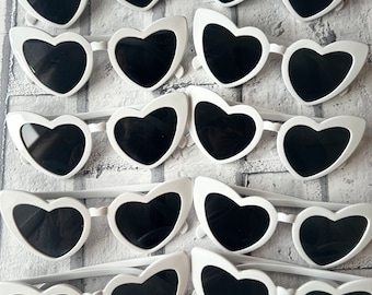 May include: A set of white heart-shaped sunglasses with black lenses. The sunglasses are arranged in rows on a white brick background.