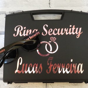 May include: Black ring security case with a rose gold inscription that reads "Ring Security" and "Lucas Ferreira". The case has a pair of black sunglasses with the words "Ring Security" and "Lucas Ferreira" printed on the arms.