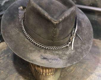 May include: A black felt fedora hat with a leather band and chain detail. The hat has a distressed look and is decorated with stitching and a feather.