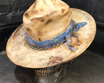 Puede incluir: Un sombrero fedora beige desgastado de ala ancha, con un pañuelo azul estampado alrededor de la copa, una cadena metálica y una pequeña llave de metal. El sombrero tiene marcas de quemaduras, detalles de costura azul y el texto FOLLOW YOUR HEART DESIRE.