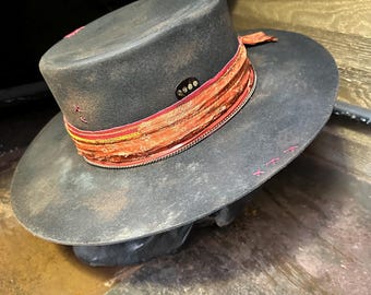 May include: A dark gray hat with a wide brim and a flat top. The hat features a decorative band in shades of orange and red, with a small, circular embellishment. Pink stitching accents the hat's crown and brim.