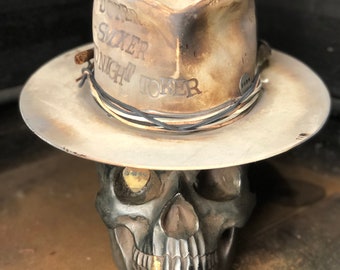 Puede incluir: Un sombrero vaquero de cuero blanco con las palabras "Joker Midnight Joker" grabadas en la corona. El sombrero está sentado encima de una calavera negra.