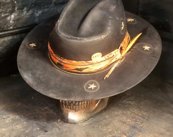 Puede incluir: Un sombrero fedora de fieltro negro con ala ancha adornada con cuatro estrellas metálicas. La copa está envuelta en una banda de tela naranja texturizada con una fina cadena dorada, un pequeño emblema metálico y una pluma insertada en el lateral.