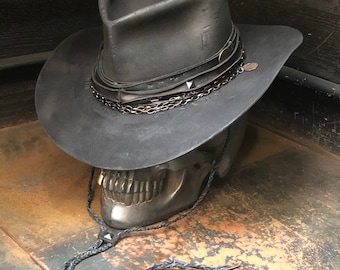 May include: A black leather cowboy hat with a wide brim and a skull-shaped base. The hat is decorated with multiple leather bands and a chain.