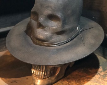 Può includere: Un cappello in feltro grigio scuro con una corona modellata a forma di teschio e una tesa larga. Il cappello presenta un sottile cordino in pelle scura avvolto attorno alla base della corona, fissato con un piccolo perno metallico. Poggia su un teschio metallico.