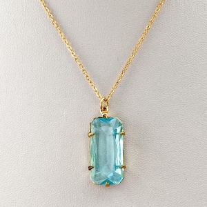 Puede incluir: Un collar dorado con un colgante rectangular de piedra preciosa azul claro. El colgante está engastado en un marco dorado y cuelga de una delicada cadena dorada. El collar se muestra sobre un fondo blanco.