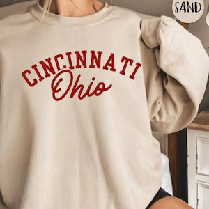 Puede incluir: Una sudadera de color arena con las palabras "CINCINNATI Ohio" impresas en rojo. El texto está arqueado sobre el pecho. La sudadera tiene mangas largas y cuello redondo.