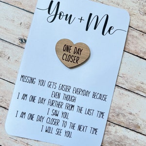 May include: A white card with the text "You + Me" in black cursive writing. A small wooden heart with the text "One Day Closer" is attached to the card.
