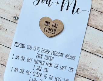 May include: A white card with the text "You + Me" in black cursive writing. A small wooden heart with the text "One Day Closer" is attached to the card.