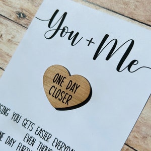 May include: A white card with black text that reads "You + Me" and a wooden heart with the text "One Day Closer".