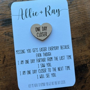 May include: A white card with the names Allie and Ray written in cursive. A wooden heart with the text "One Day Closer" is attached to the card. The card has a message about missing someone and being one day closer to seeing them again.