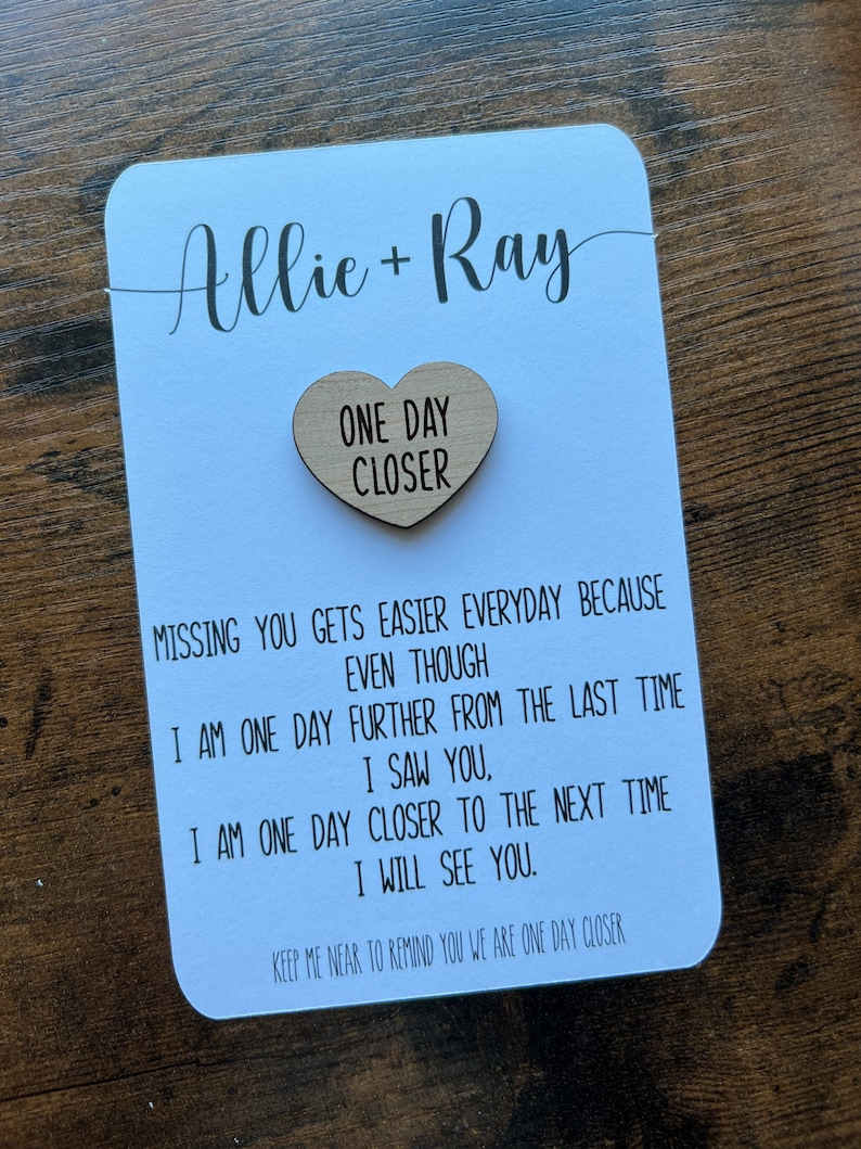 May include: A white card with the names Allie and Ray written in cursive. A wooden heart with the text "One Day Closer" is attached to the card. The card has a message about missing someone and being one day closer to seeing them again.