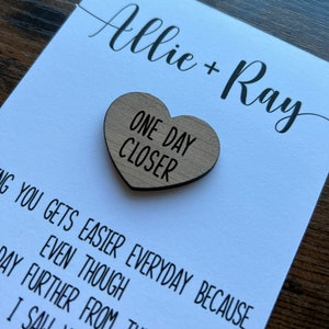 May include: A white card with the names Allie + Ray written in black cursive. A wooden heart with the text "One Day Closer" is attached to the card.