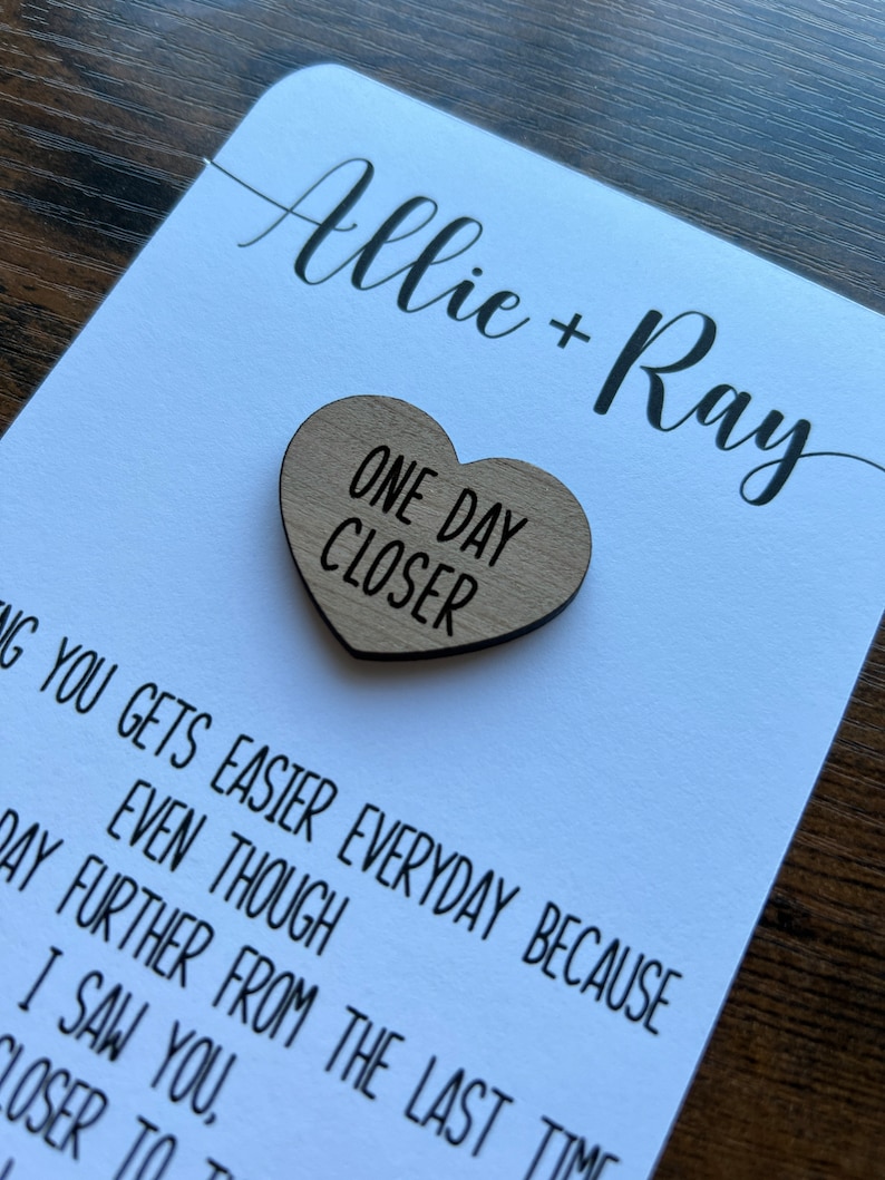 May include: A white card with the names Allie + Ray written in black cursive. A wooden heart with the text "One Day Closer" is attached to the card.