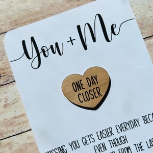 May include: A white card with black calligraphy that reads "You + Me" and a small wooden heart with the text "One Day Closer" in black lettering.