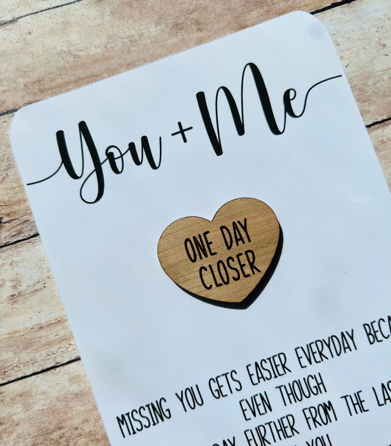 May include: A white card with black calligraphy that reads "You + Me" and a small wooden heart with the text "One Day Closer" in black lettering.