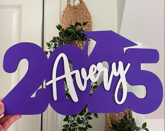 May include: Purple wooden sign with the year 2023 and the name Avery in white script. The sign is shaped like a graduation cap.