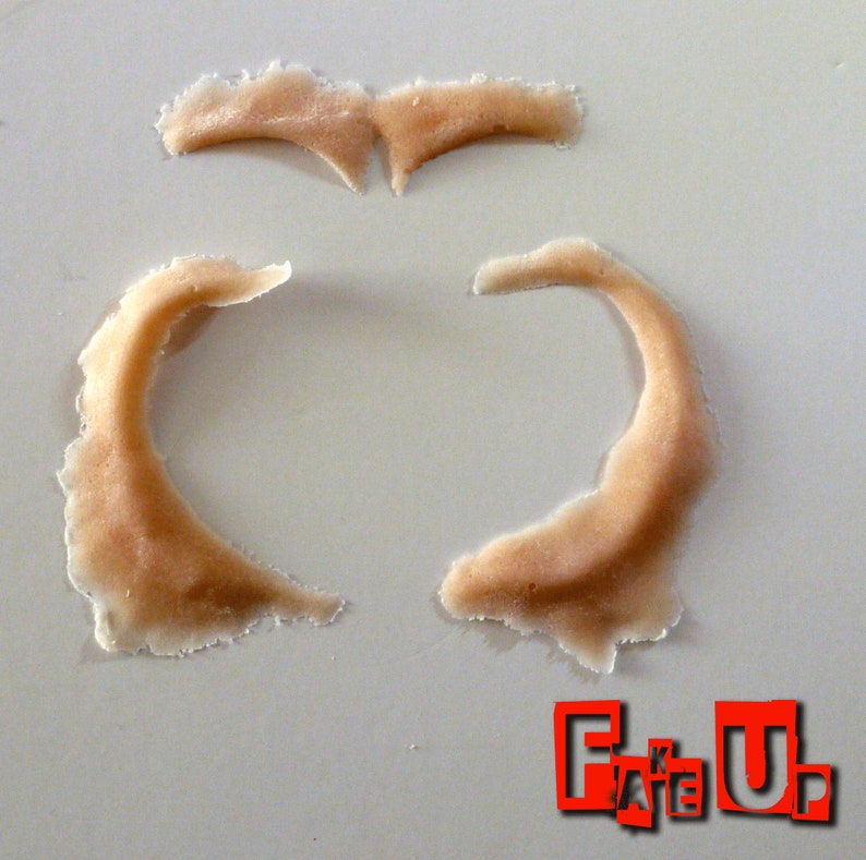 May include: Three pieces of flesh-toned silicone prosthetic makeup. The pieces are shaped like eyebrows and a pair of ears.