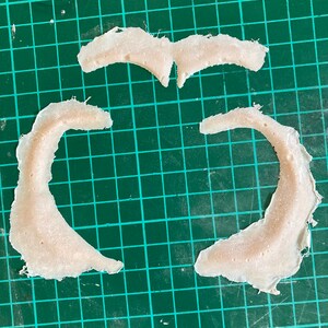 May include: Three pieces of white, textured, fabric cut into curved shapes. The fabric is laid out on a green cutting mat.