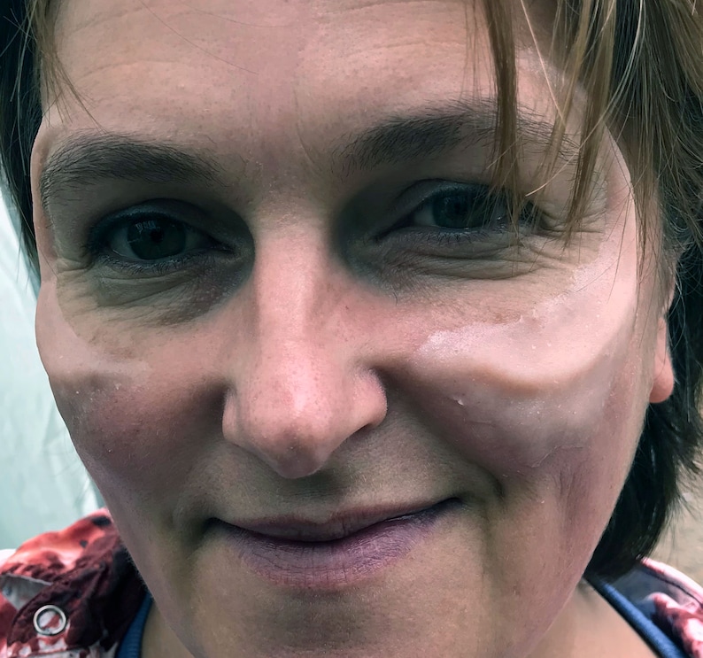 May include: Close-up of a woman's face with a light-colored cream or makeup applied to one cheek. The woman is smiling and has brown hair.