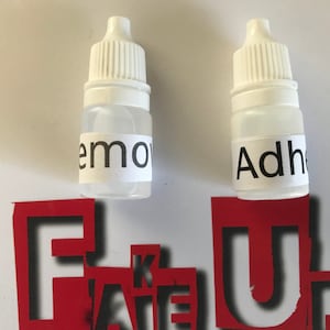 Puede incluir: Dos pequeños frascos blancos con goteros. El frasco de la izquierda tiene una etiqueta que dice "Remove" y el frasco de la derecha tiene una etiqueta que dice "Adhesive".