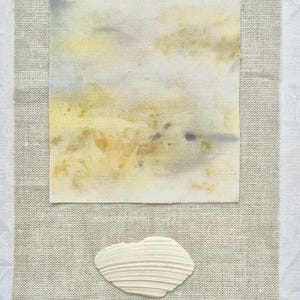 May include: A square watercolor painting with yellow, brown, and blue hues, resting on a beige linen fabric. A white seashell is positioned below the painting. The background is a white, patterned surface.