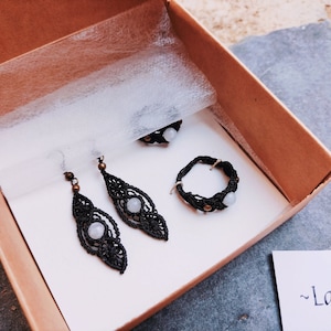 May include: A set of three jewelry pieces in a brown box. The set includes a ring, a pair of earrings, and a necklace. All of the pieces are made with black thread and feature a white stone.