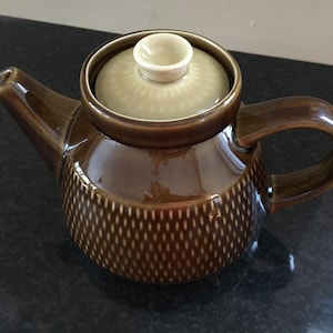 Puede incluir: Una tetera de cerámica marrón con una tapa marrón claro y un diseño estampado. La tetera tiene un cuerpo redondo y un asa curva.