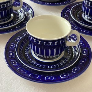 Könnte beinhalten: Ein Satz mit vier blauen und weißen Keramik-Teetassen und Untertassen mit geometrischem Muster. Die Tassen und Untertassen sind in einem Kreis auf einer weißen Tischdecke angeordnet.