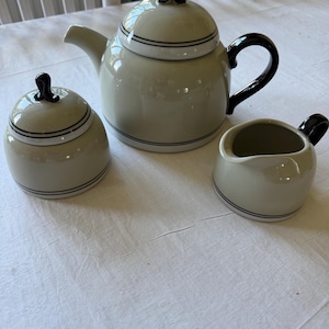 Puede incluir: Un juego de té de cerámica que consta de una tetera, un azucarero con tapa y una jarra para leche. Cada pieza es de color beige claro con acabado brillante, con finas rayas horizontales negras y asas y remates de tapa en color negro.